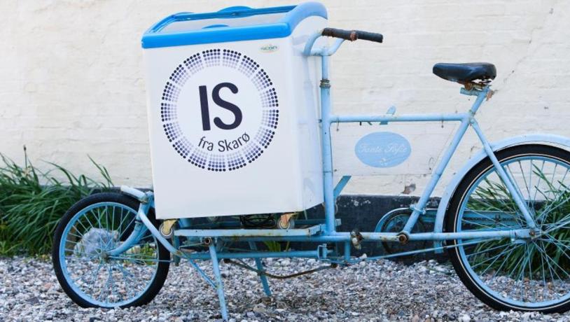 Lyseblå is-cykel med logoet "Is fra Skarø" står parkeret ved en væg.