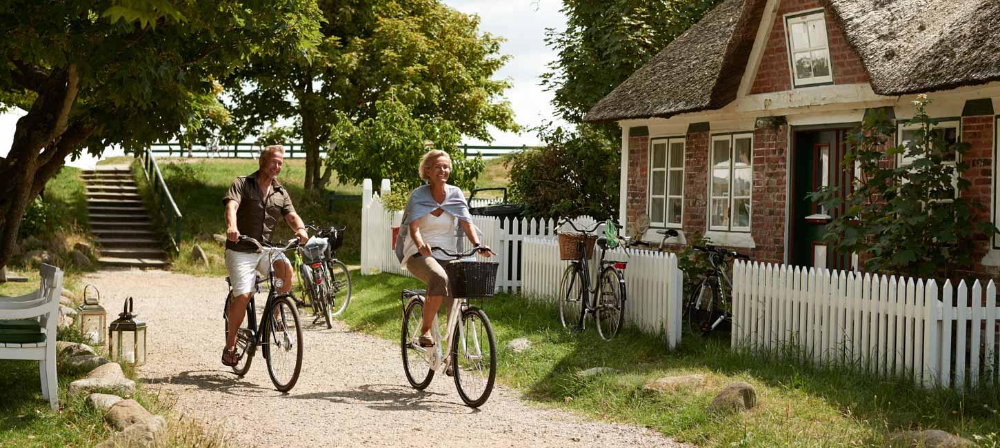 Et par cykler igennem idyllisk landskab med stråtagshus, hvidt stakit og grønne træer i baggrunden.