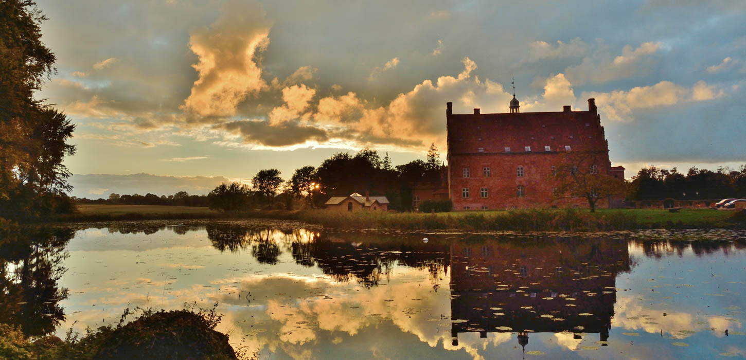 Spejling af det smukke røde slotshotel Broholm Slot i sø med oplyste skyer som baggrund.