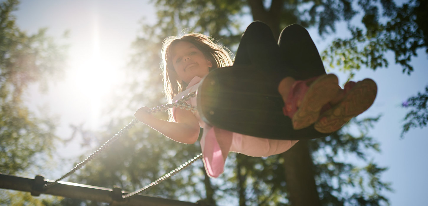 Pige, der gynger, set nedefra og i modlys.. Hun kigger ned og smiler stort.