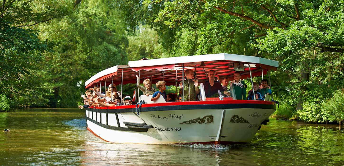 Mange mennesker er ude at sejle med Odense Aafart på en solskinsdag. Båden er hvid og rød og træerne omkring er grønne.