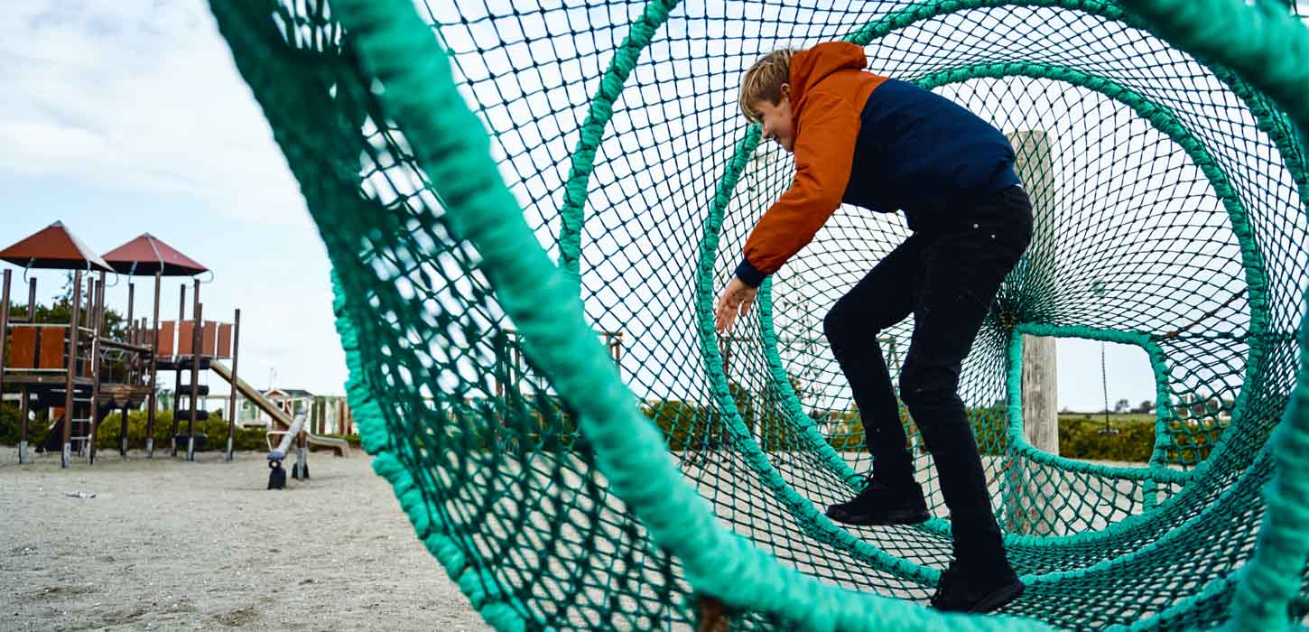 Et større barn inde i 'fiskeruse' på en legeplads med faldsand. I baggrunden ses et legetårn.