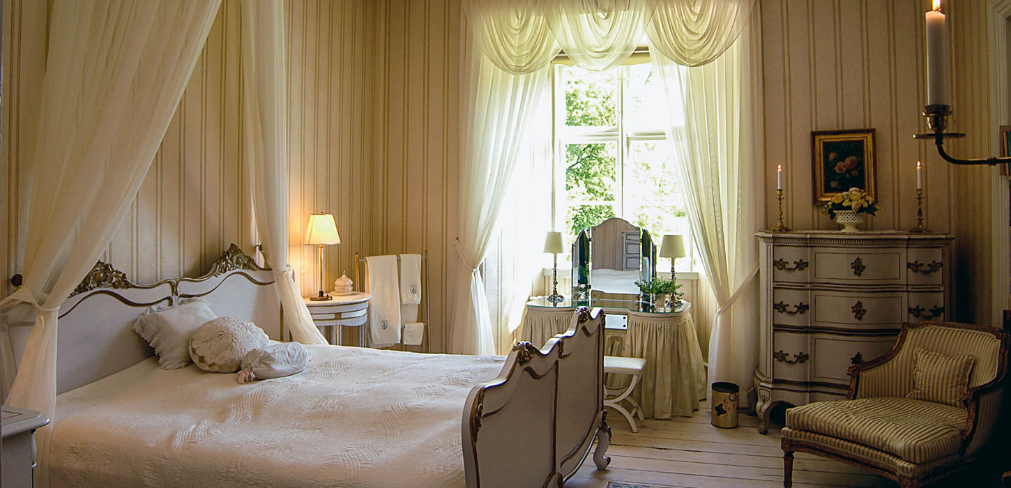 Suite med smuk, gammel seng, oplyste hvide gardiner og andet gammelt interiør på på Broholm Slot.