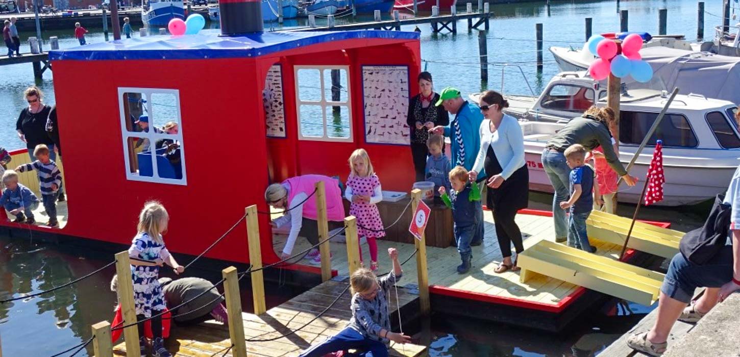 En rød åben husbåd er lavet om til en legeplads. Den er fyldt op med legende børn og opmærksomme forældre. I baggrunden ses et par almindelige både tøjret til havnen.