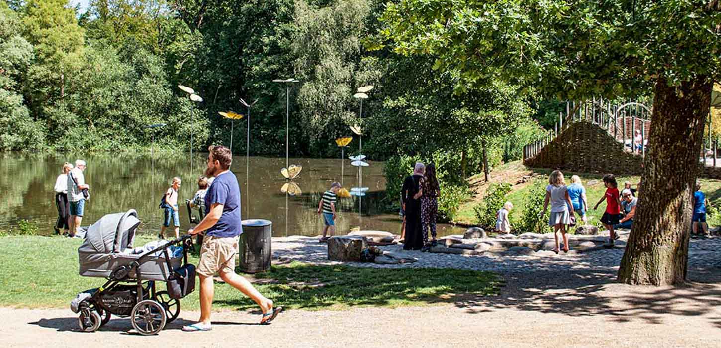 Mand med barnevogn går forbi børn, der leger ved vand med store kunstsommerfugle på pinde.
