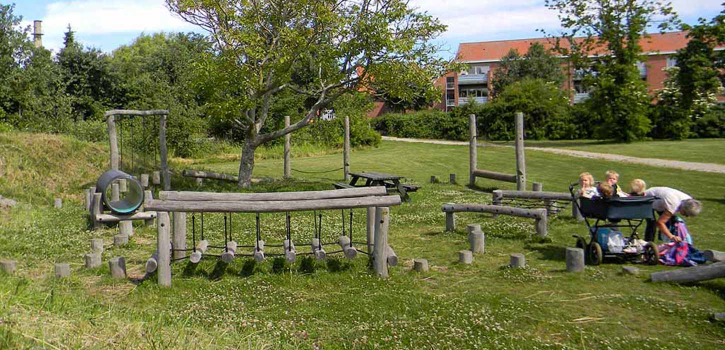 På en græsplæne ved siden af en lille sti er en legeplads lavet af træ. I højre side står en barnevogn med fire børn og en kvinde ved siden af. 