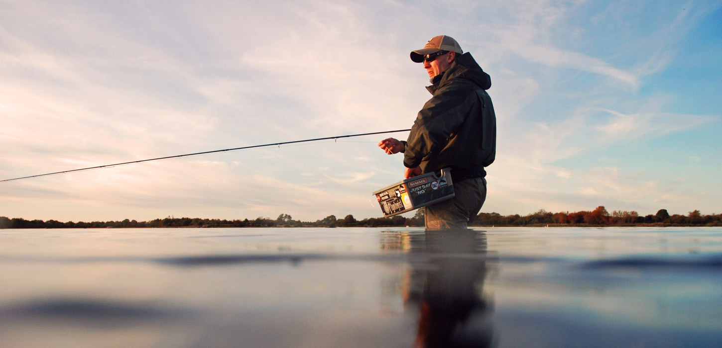 Fisker står i vandet i vaders og fiskeudstyr og fisker. Vandet er spejlblankt og himlen er blå, med luftige, hvide skyer.