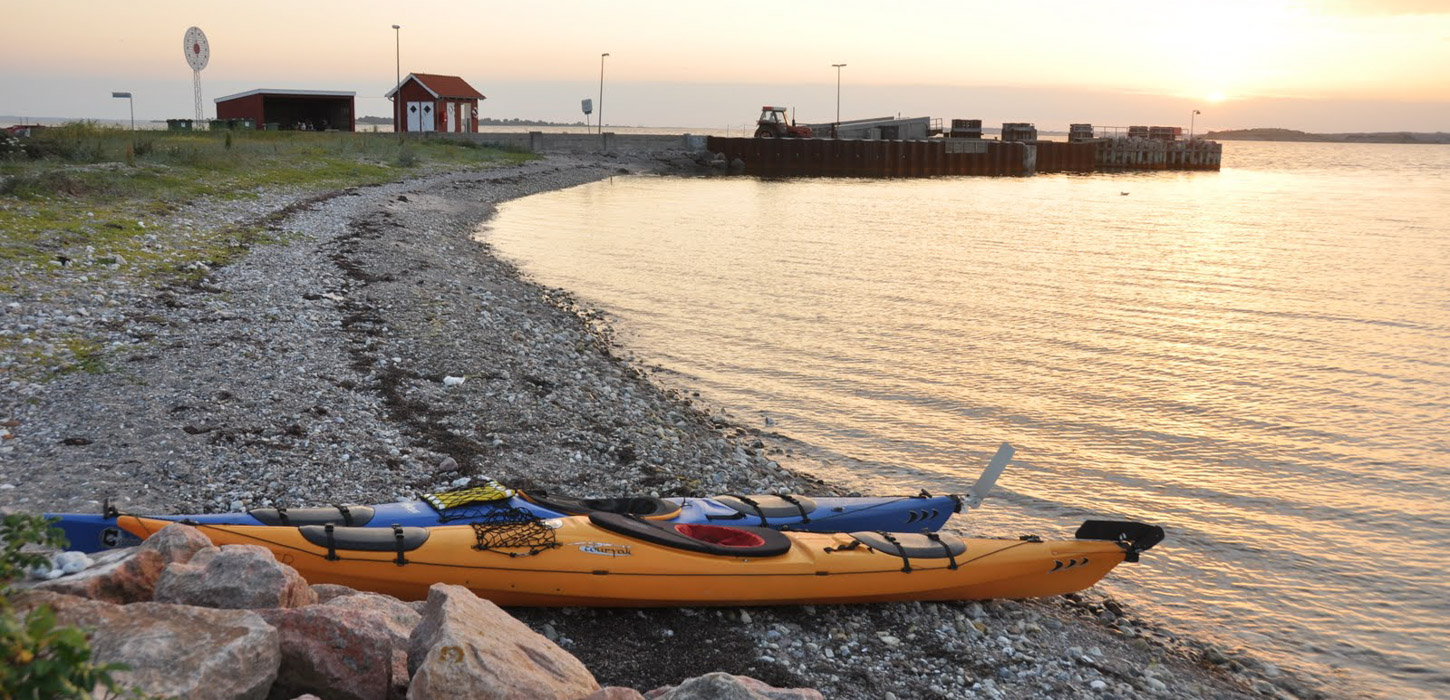 På billedet ses en strandbred. Den fylder hele venstre side af billedet og ender i baggrunden af billedet. Her står to små røde bygninger. Henover strandbredden er solen på vej ned. I højre side er havet. i forgrunden ligger en gul og en blå kajak på standen.