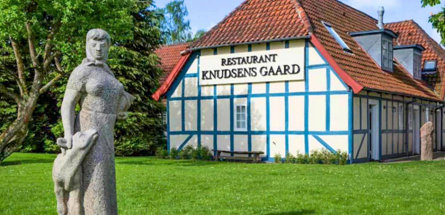 Stenstatue, der forestiller en kvinde og en kalv på en grøn græsplæne foran en bindingsværksbygning med blåt bindingsværk og rødt tegltag.