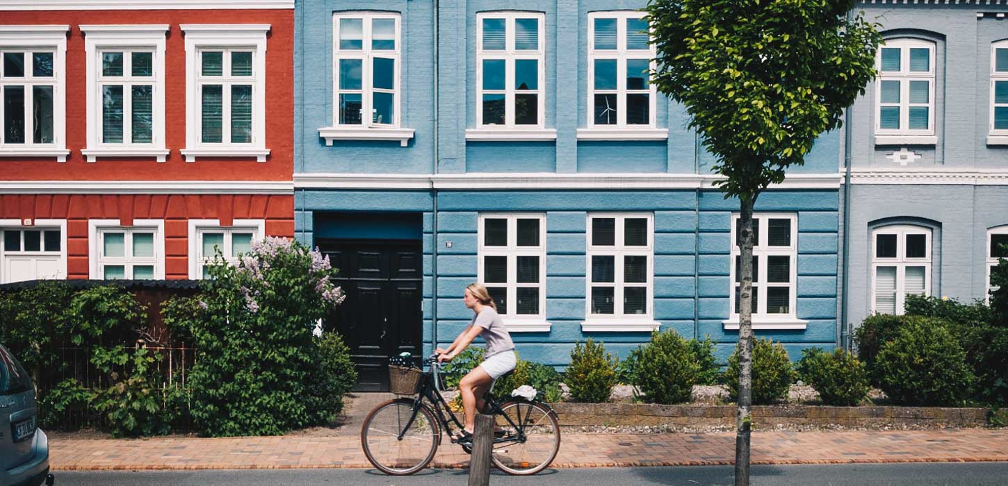 Kvindelig cyklist foran en række af byhuse; et rødt, et blåt og et gråt.