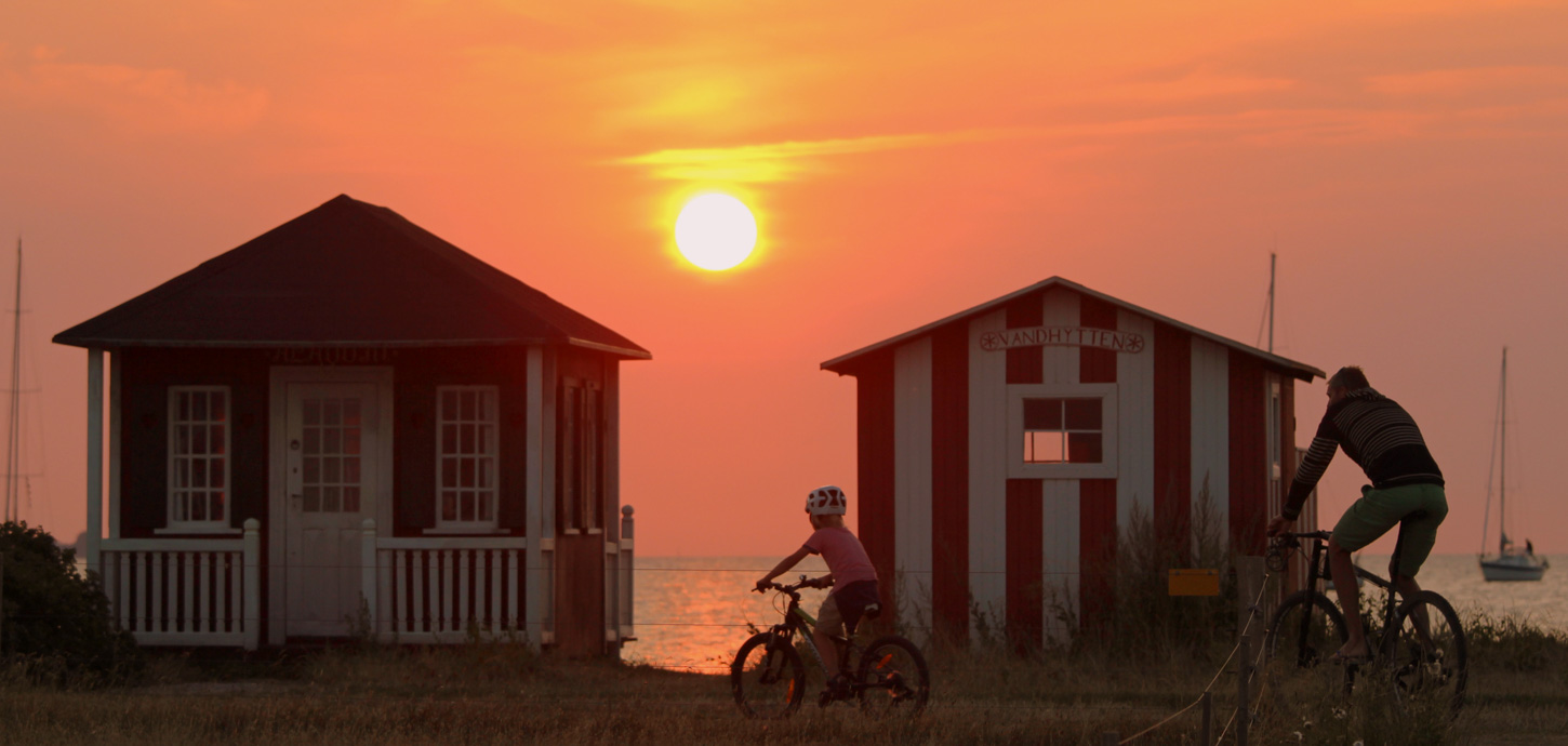 Far og barn på cykel i knaldrød solnedgang foran to små badehuse på Ærø.