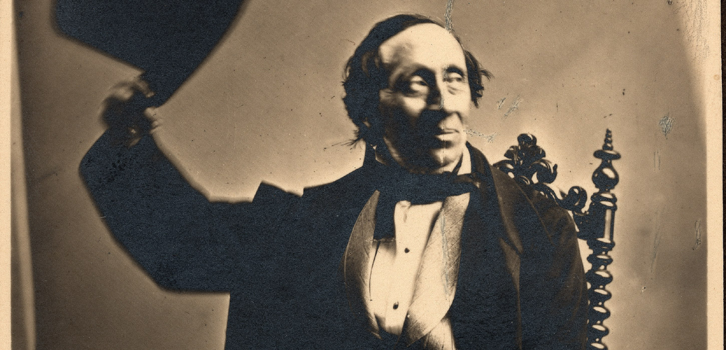 H. C. Andersen sidder på stol og løfter sin hat