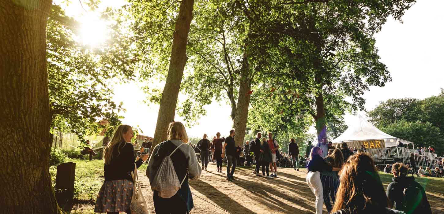 På en grussti under flere træer går en masse mennesker op imod de plastiktelte der står i baggrunden. 