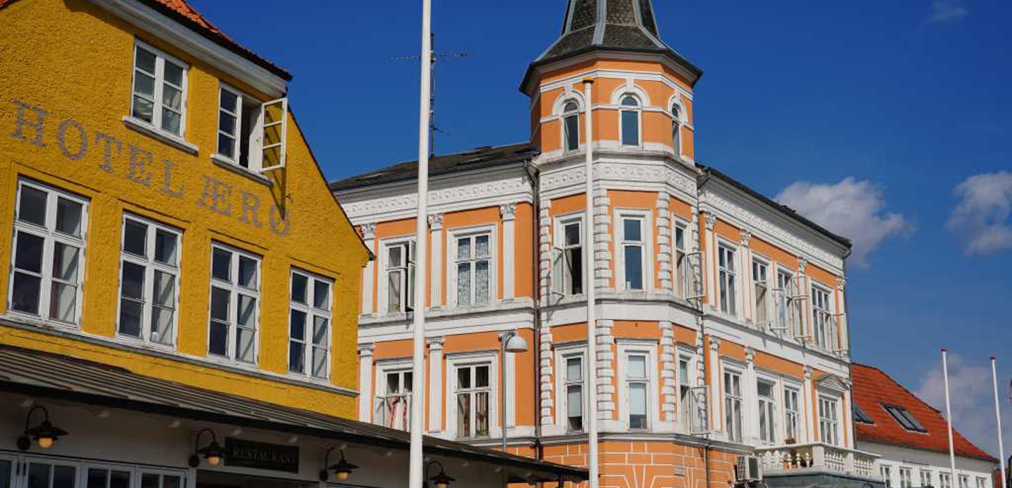 I venstre side ses toppen af en sennepsgul bygning med teksten Hotel Ærø. I højre side ses en orange bygning med hvide vinduesrammer og et tårn.