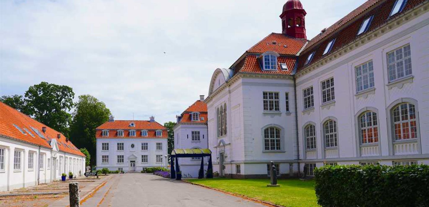 Stor, hvid herregård med rødt tag og en masse vinduer, en vej og en grøn forhave.