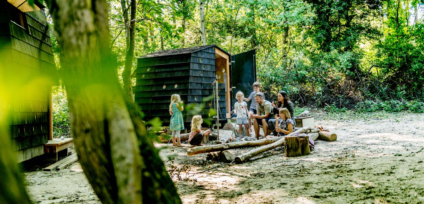 Fem børn og to voksne samlet om et bål i skoven. Bag ved dem ses et arkitekttegnet shelter.