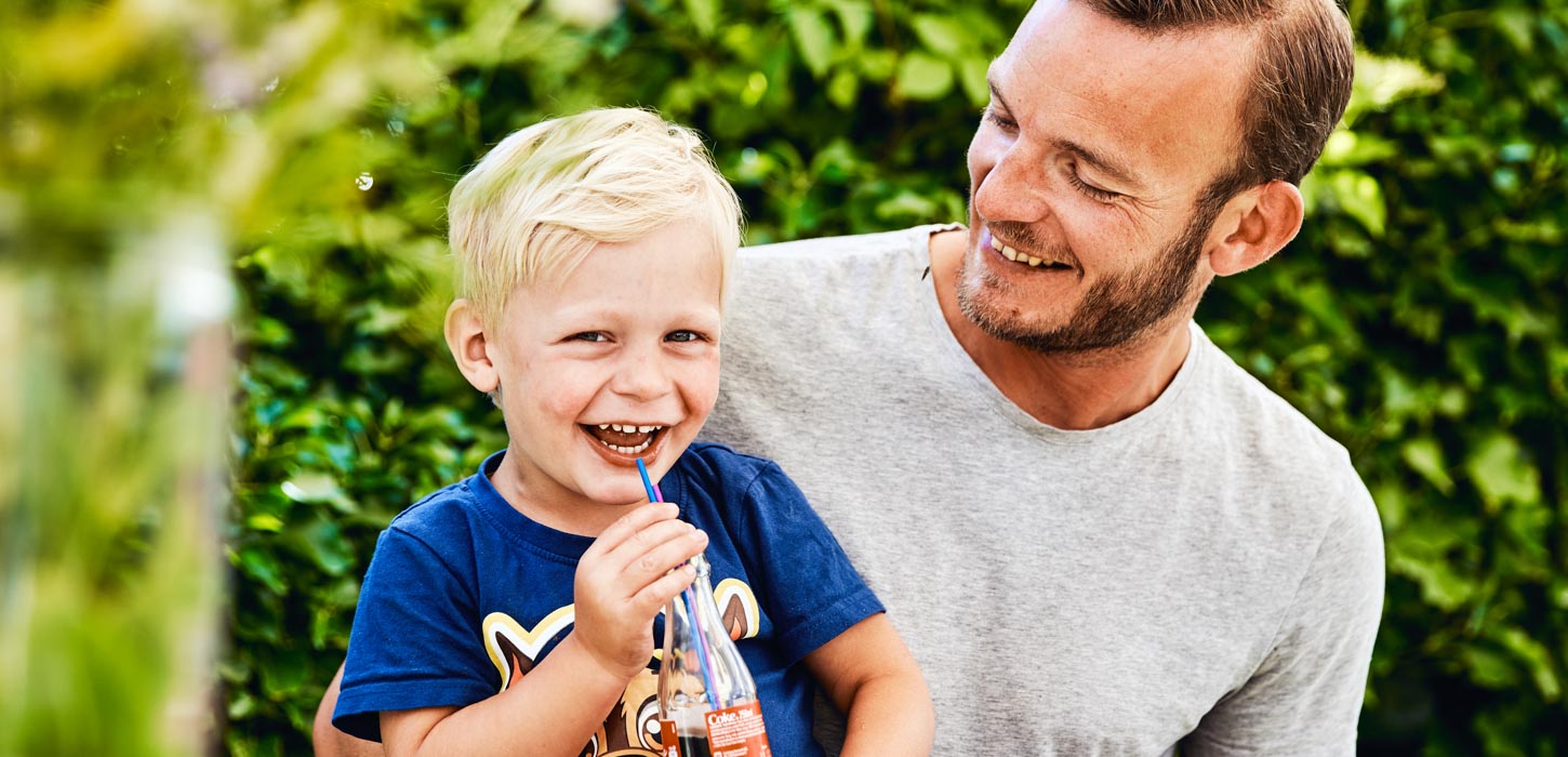 En lille lyshåret dreng sidder på skødet af sin far. Drengen har en blå t-shirt på og en cola med sugerør i hånden. Han smiler stort til kameraet, men hans far kigger smilende ned på ham. De er omgivet er en grøn hæk.