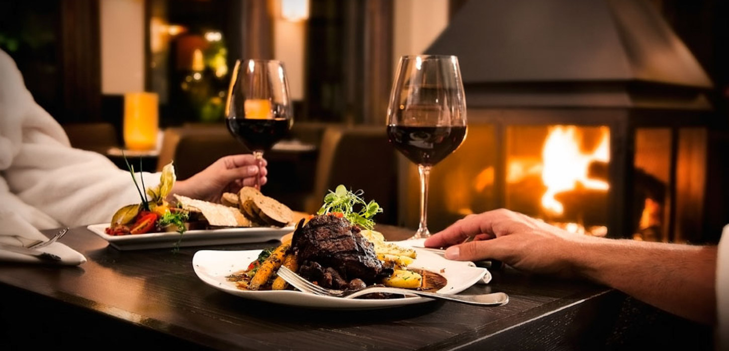 Et nært billede af et sort topersoners restaurantbord. På bordet står to tallerkner med bøf, sovs og grøntsager. To hænder holder hver deres glas vin. I baggrunden brænder en stor sort pejs. 