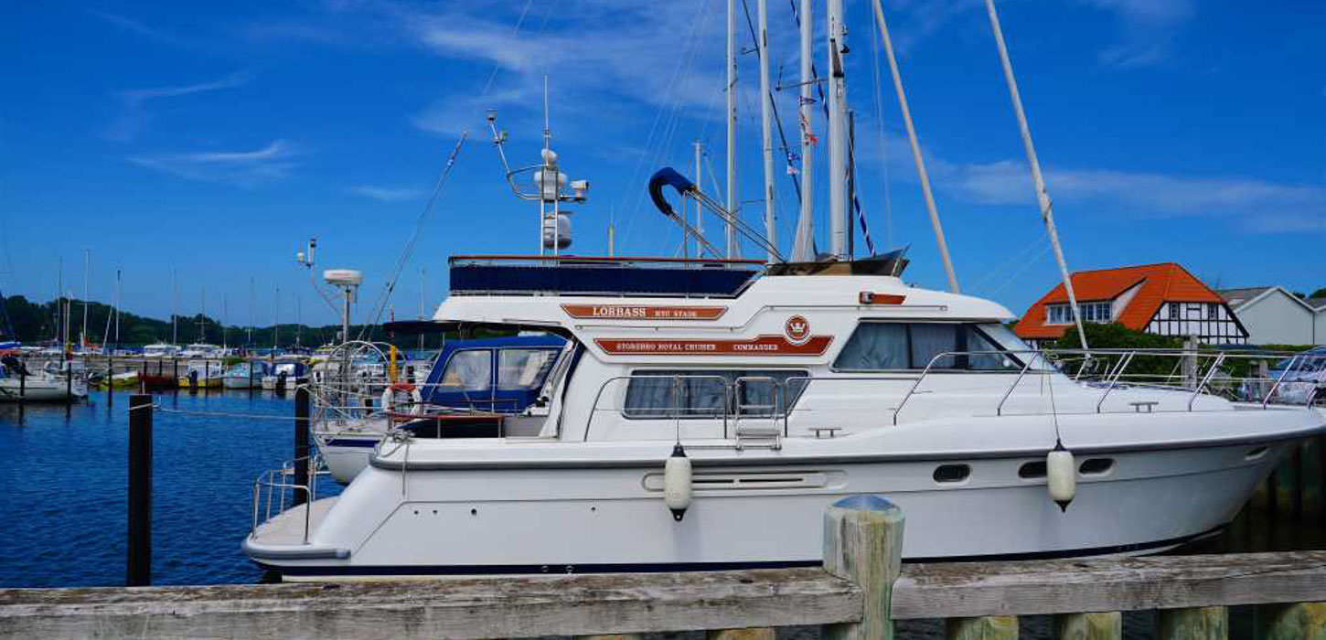 Under hel blå himmel ses en hvid speedbåd. Båden ligger til kaj i en havn, og i baggrunden kan man se mange flere forskellige både. Vandet er helt blåt, og i højre side af billedet ses en hvidt bindingsværkshus med rødt tag.