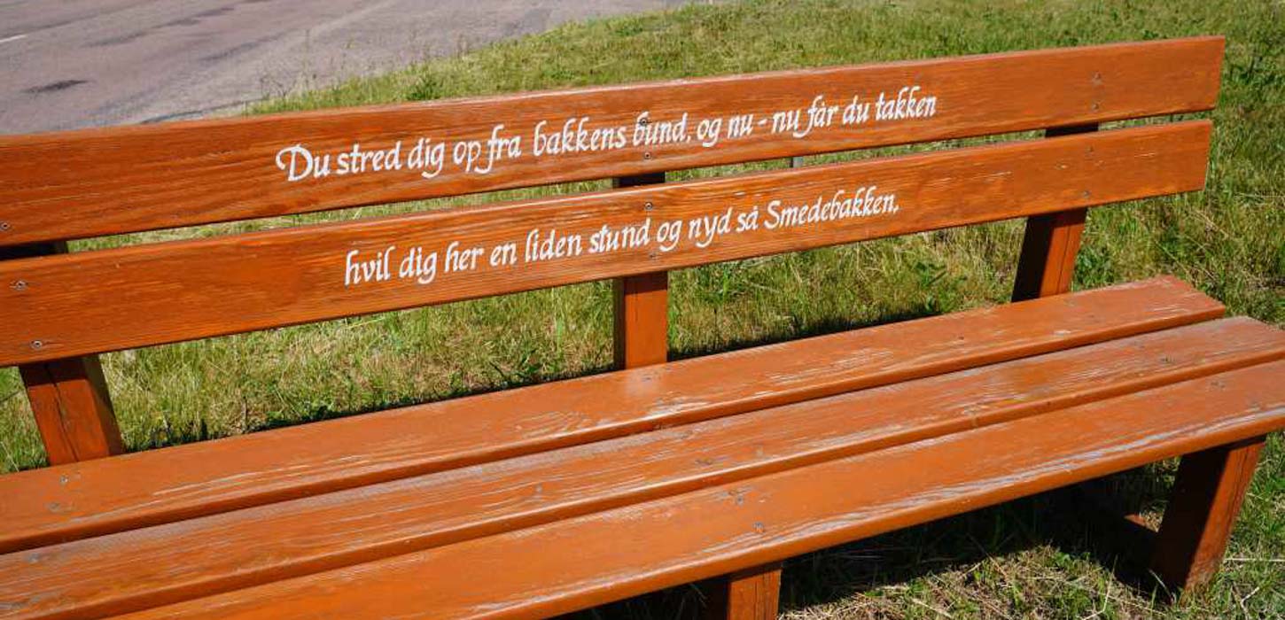 Nærbillede af brun træbænk med teksten: Du stred dig op fra bakkens bund, og nu - nu får du takken. Hvil dig her en liden stund og nyd så Smedebakken.