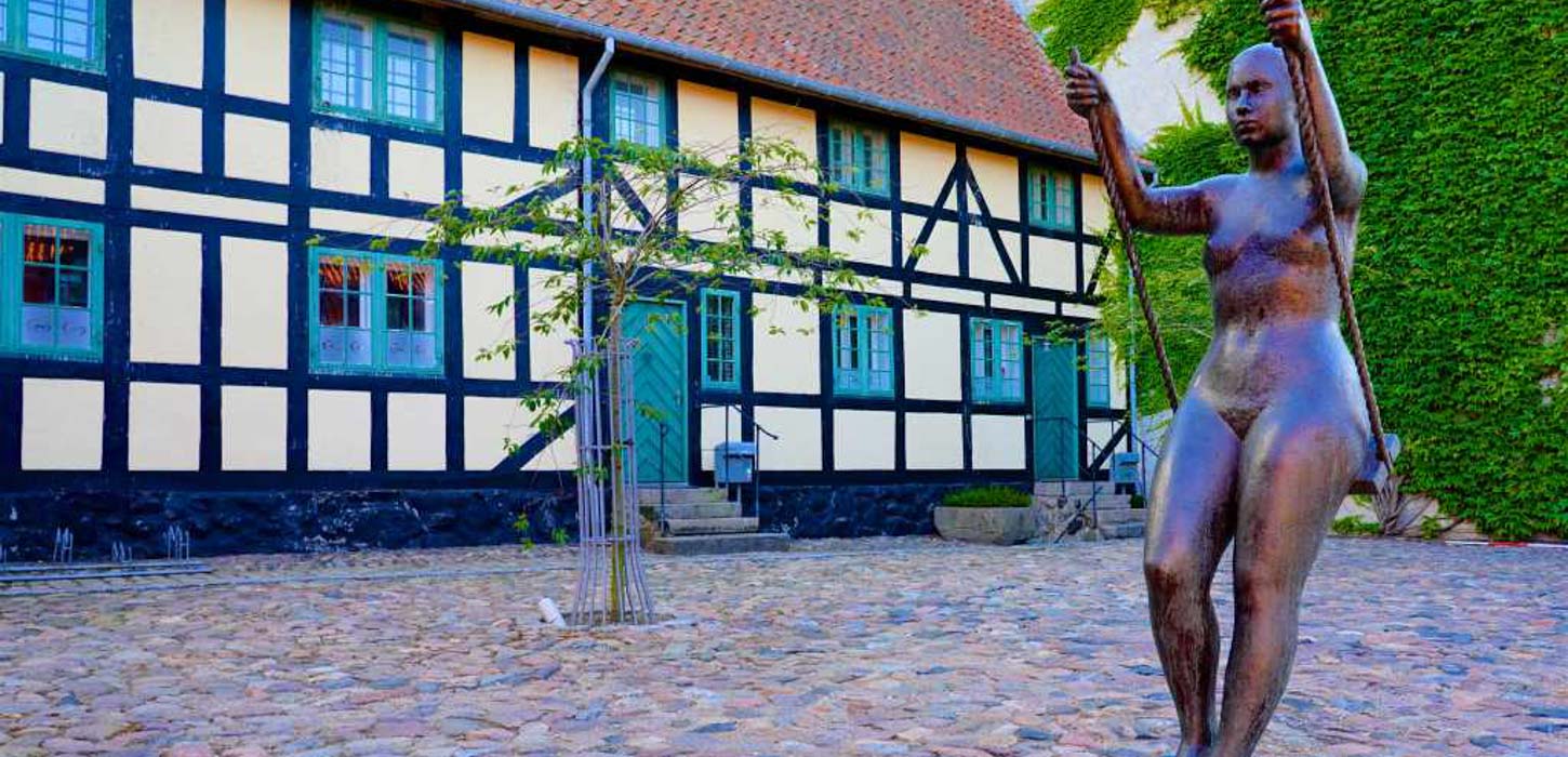 En statue af en nøgen kvinde, der holder i rebene på en gynge, på en brostensbelagt plads foran en traditionel bindingsværksbygning. Der er et ungt træ med en støttepæl ved foran bygningen,.