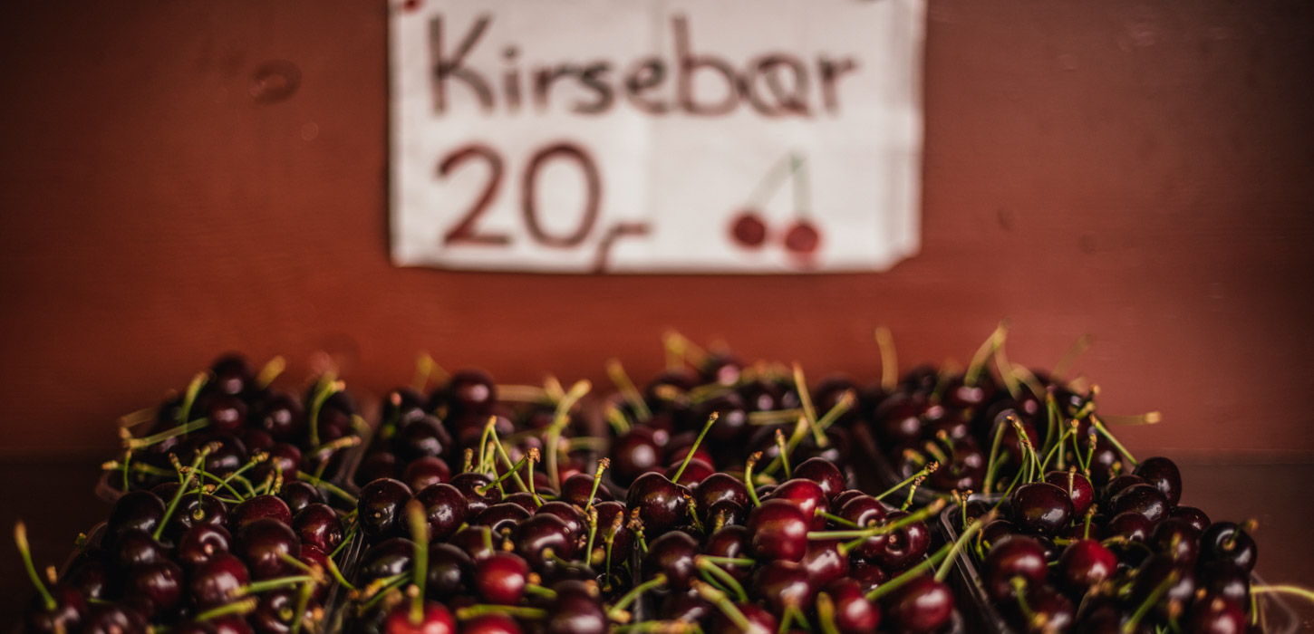 Otte pap-bakker er fyldt med kirsebær. De står på en træplade. Bagved er en sløret rød væg, med et hvidt papskilt. På skiltet står der "Kirsebær 20kr", og der er tegnet et kirsebær.