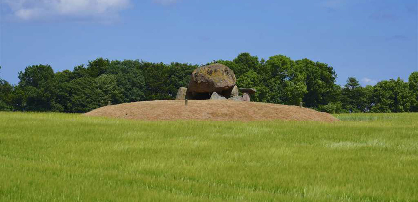 Midt på en grøn mark er en lille ophøjning med en bunke sten. I bagrunden er der skov.