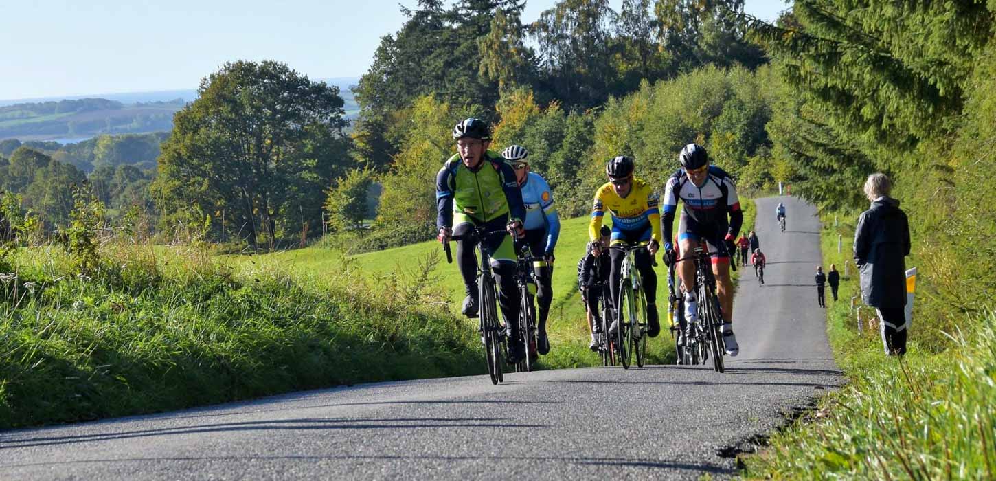 Motionscykelryttere på vej op ad lille bakke. En tilskuer står i vejkanten.