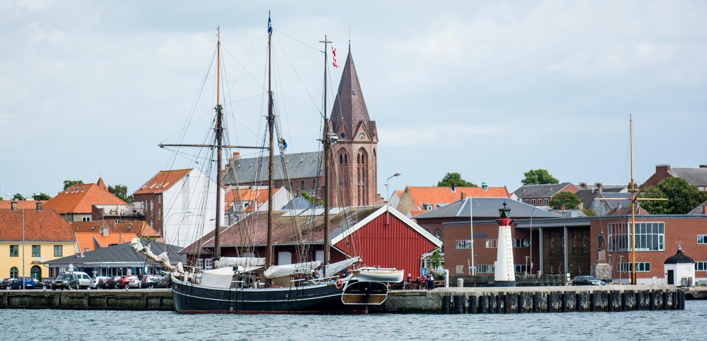 Rustikt tre-mastet træskib, der ligger til kaj med Assens by og rødstenskirken Vor Frue Kirke i baggrunden