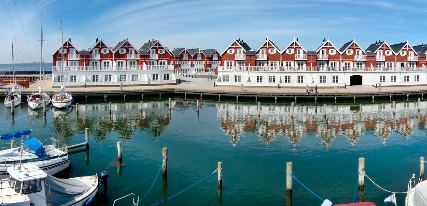 Udsigt fra en bådebro ud over vand, hvor en række end røde og hvide huse spejles fra den modsatte kant.