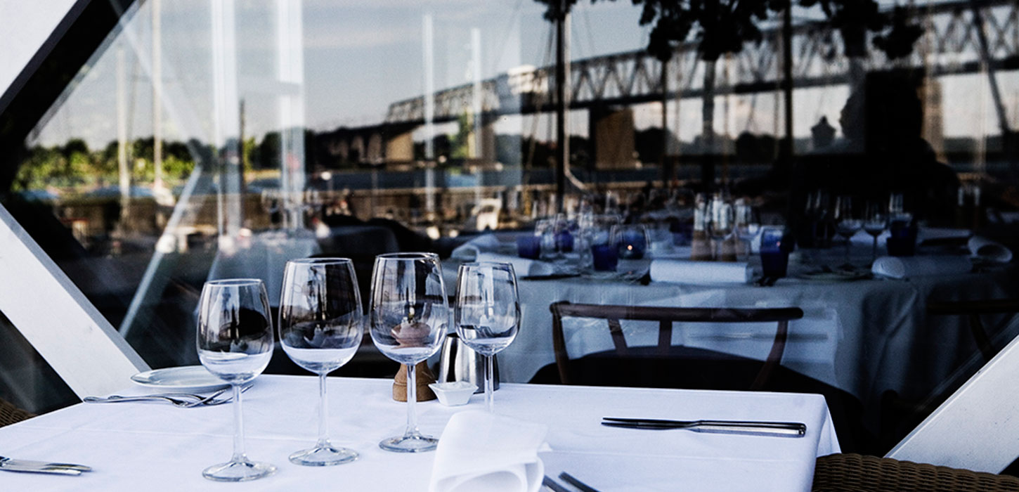 Nærbillede af smukt dækket bord med hvid dug, bestil, serviet og fire vinglas på række. I spejlbilledet i vinduesruden kan man se den gamle Lillebæltsbro.