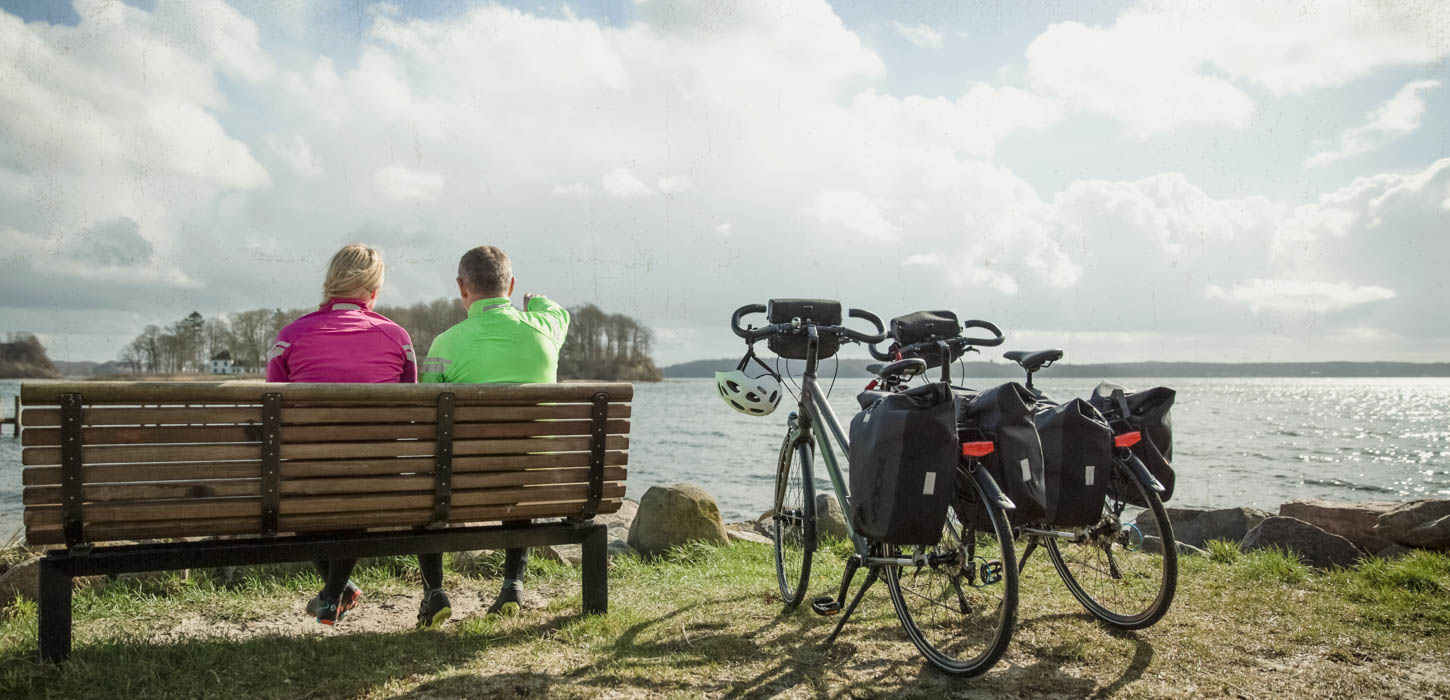 To cyklister i neon-arvede sportsjakker sidder med ryggen til på en bænk og ser ud over sundet. To cykler med cykeltasker på står parkeret ved siden af.