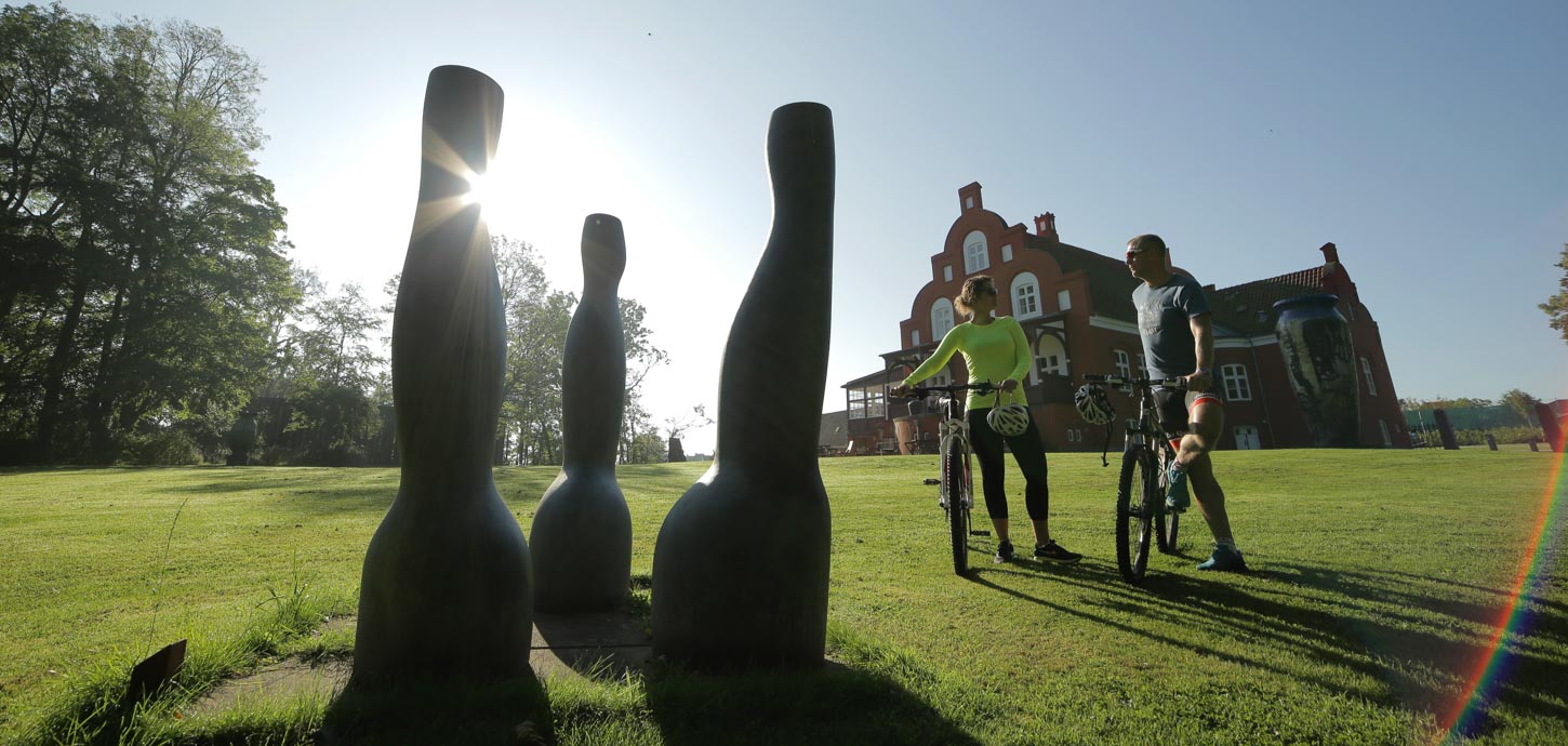 To cyklister står med deres cykler foran skulptur i solskinnet på en grøn græsplæne med en prægtig rød bygning i baggrunden.