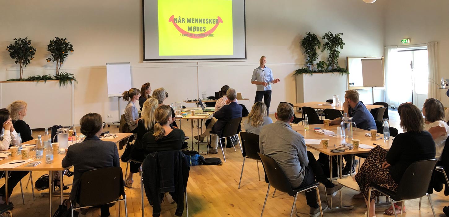 En del mennesker, siddende omkring borde med ryggen til, kigger på på oplægsholder, der er i gang med en PowerPoint-præsentation. 