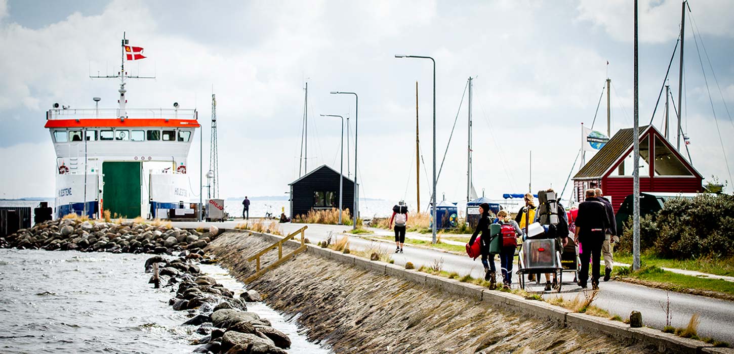 Færge ved færgeleje. Mennesker til fods med masser af oppakning går på vejen i retning mod færgen.