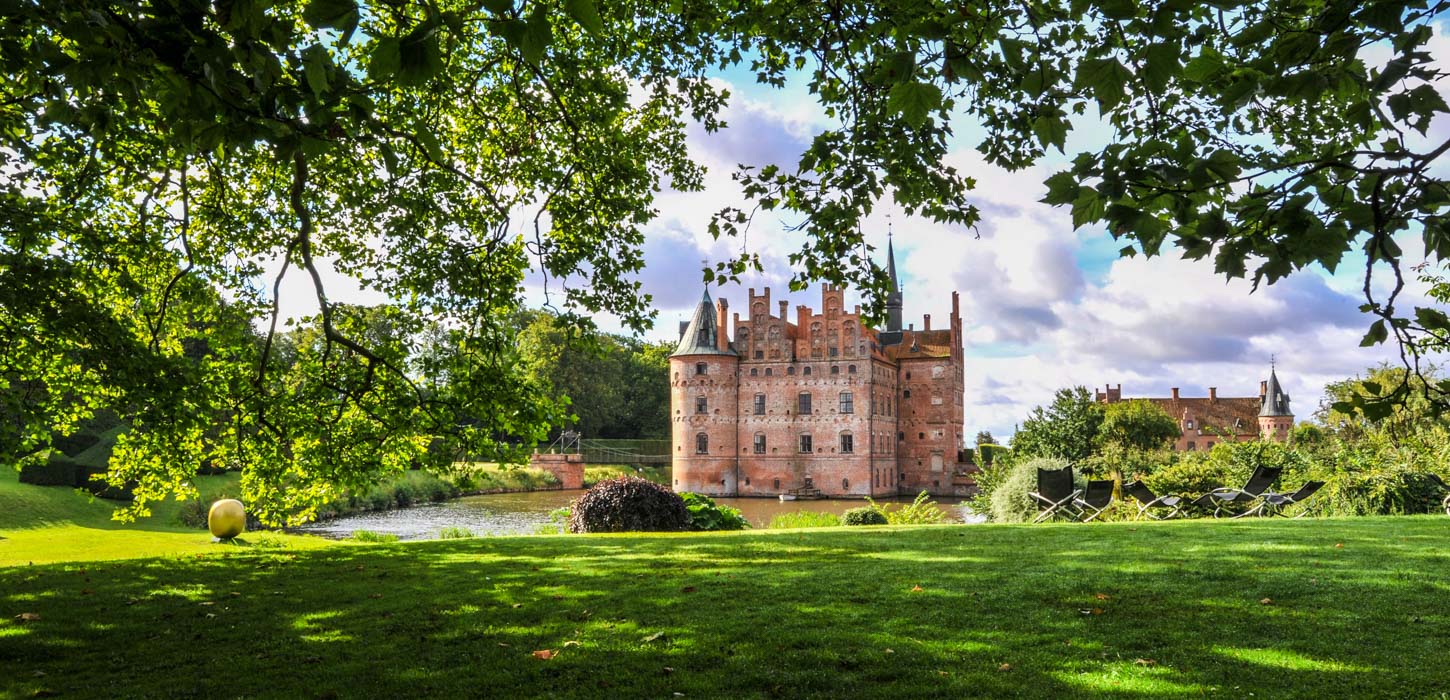 Egeskov slot med grønt græs i forgrunden og grønne træer og blå himmel i baggrunden.