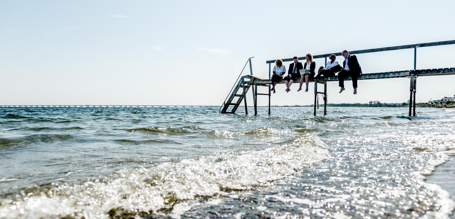 En lille gruppe forretningsfolk sidder på en badebro og kigger ud over vandet.