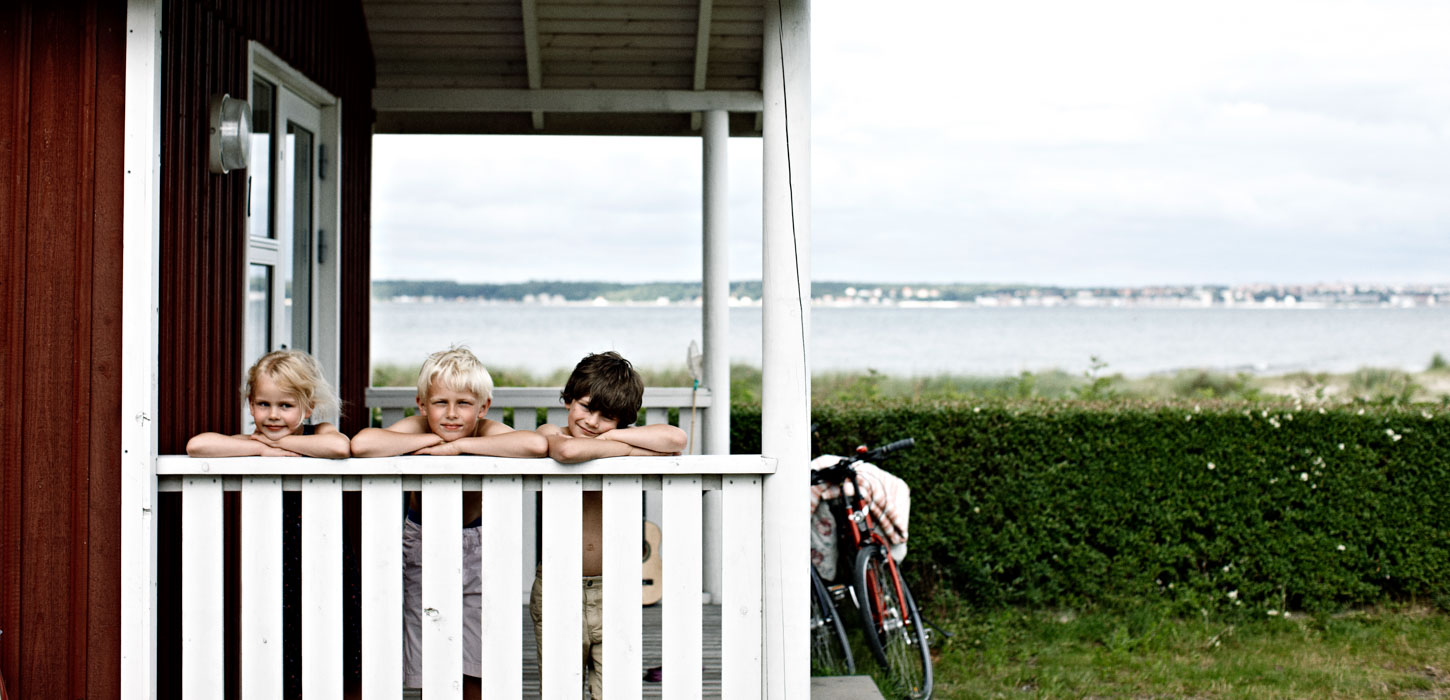 Tre børn titter op over et hvidt stakit i et sommerhus med udsigt ud til havet.