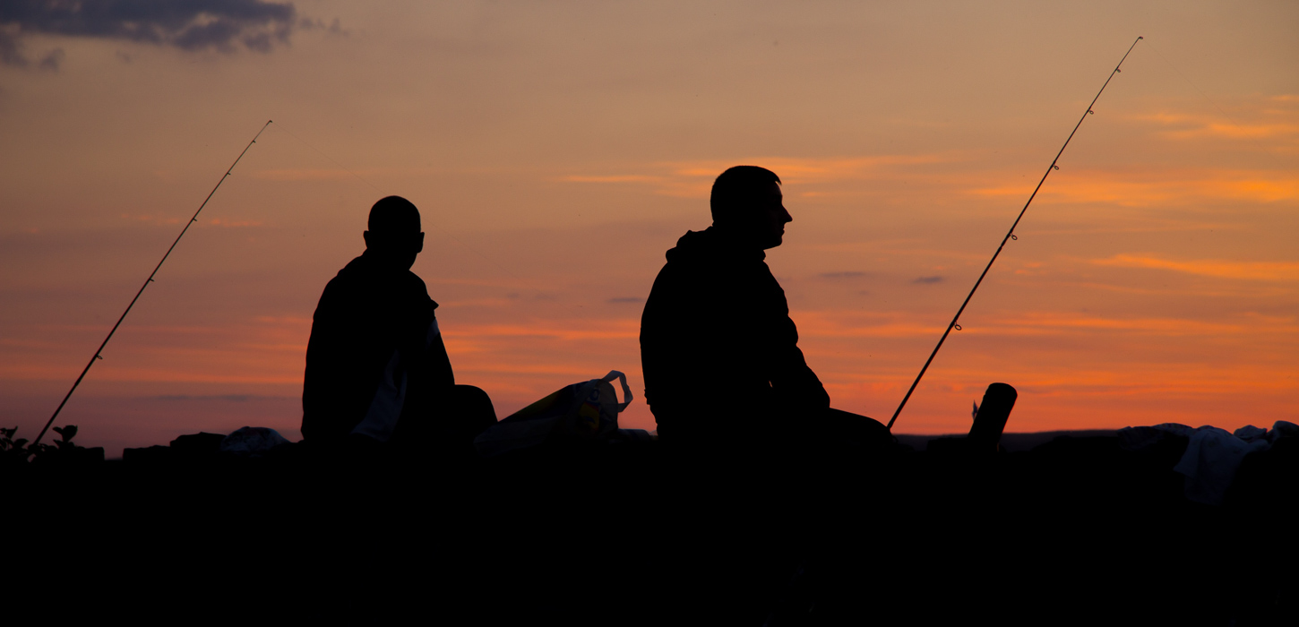 Sorte silhuetter af to skikkelser, der sidder i solnedgangen med deres fiskestænger og ser ud i horisonten.