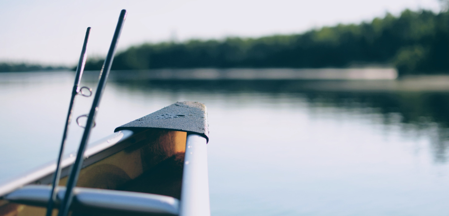Nærbillede af en fiskestang, der er delt i to. Den stikker op fra forsnuden på en kajak og i baggrunden ses det blikstille vand tæt på kysten.