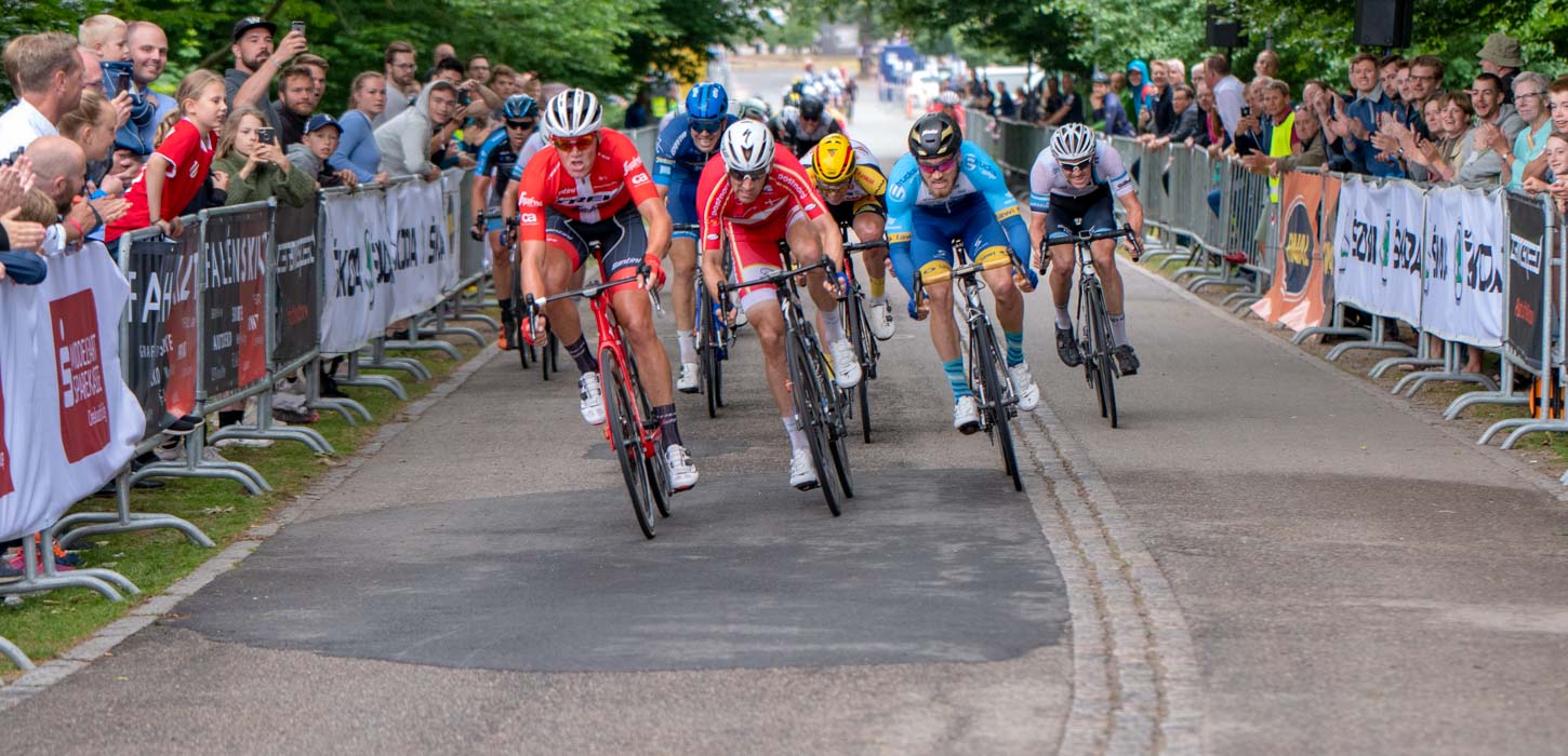 Cykelryttere på vej ind ad opløbsstrækningen i tæt løb. Tilskuere står bag hegn og hepper til begge sider.