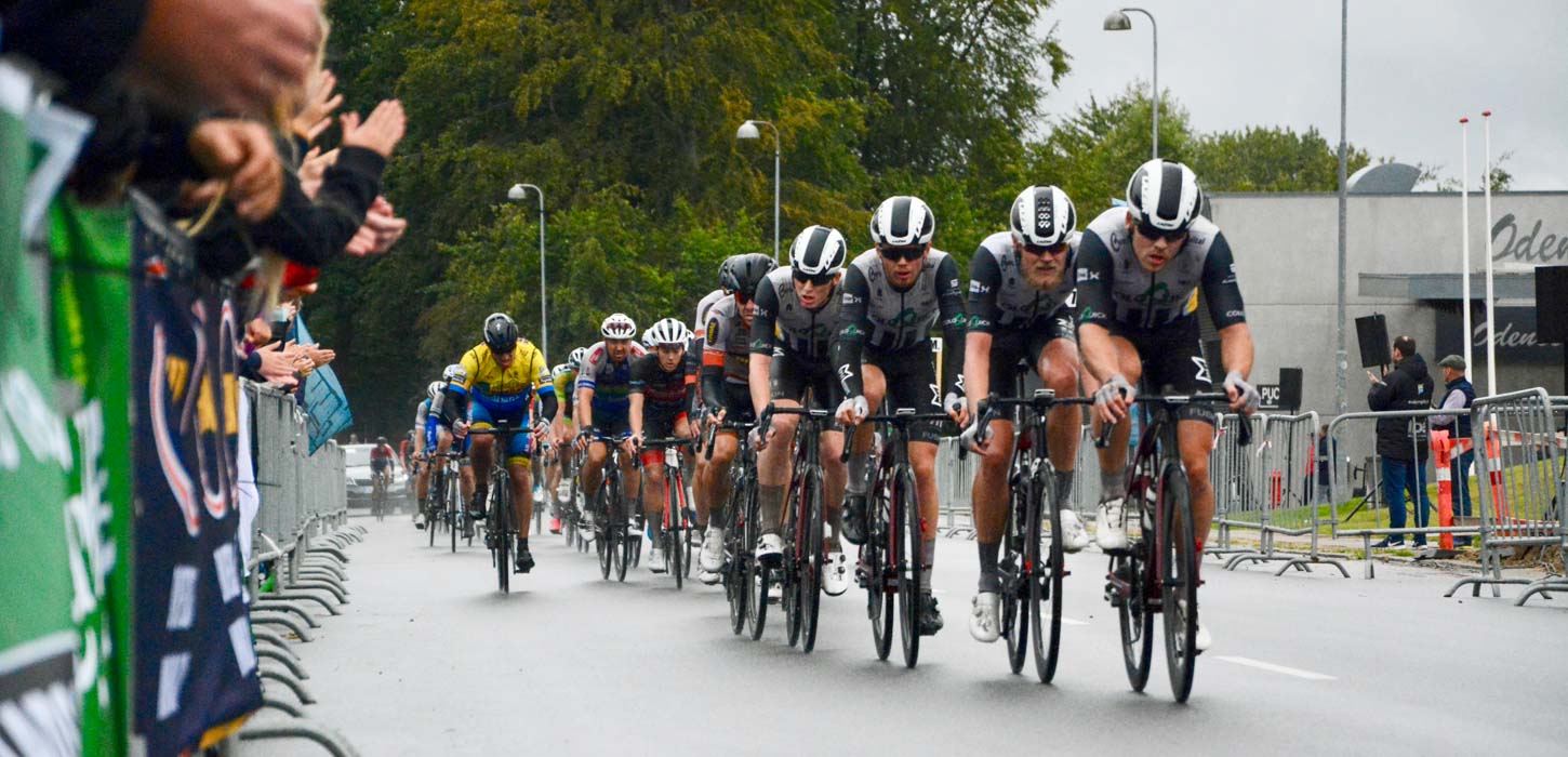 En flok cykelryttere kommer kørende ad asfaltsti langs med en afspærring med tilskuere bag.