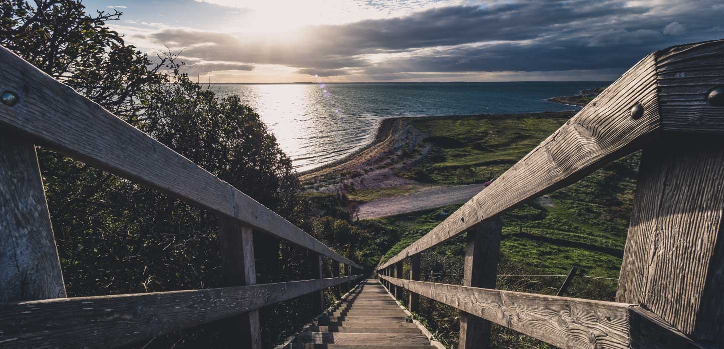 Udsigt fra toppen af lang trætrappe udover havet, som solen oplyser på dramatisk vis.
