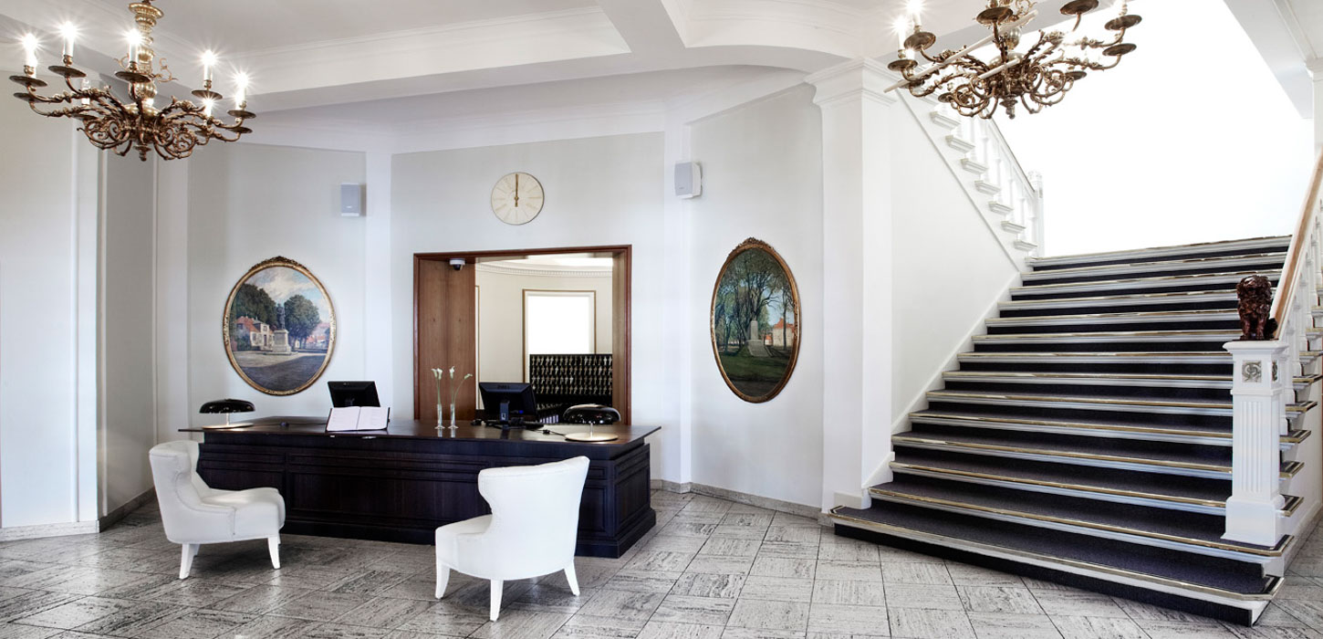 Stilren hotelreception med mork disk, hvide vægge, store hvide lænestole og runde malerier på væggene ved siden af en bred, mørk trappe.