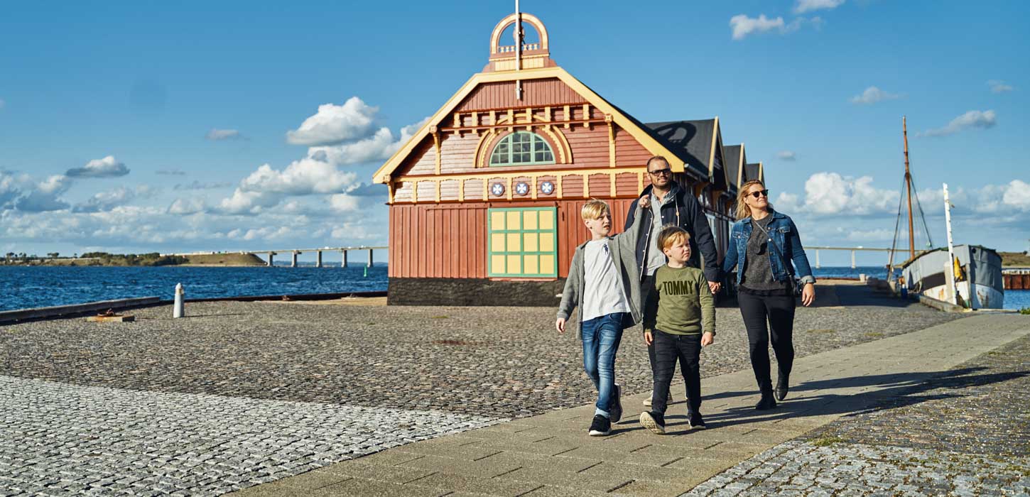 To børn og to voksne går tur ved en havn, hvor man bagved kan se et pakhus i gule og røde farver. 