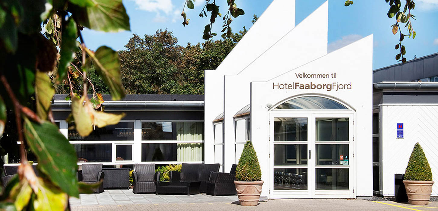 Indgangspartiet til Hotel Faaborg Fjord med en moderne hvid facade og en glasdør. Hotellets navn er fremhævet på bygningens front. Foran hotellet er en terrasse med sorte loungemøbler og to toptrimmede buske i krukker på hver side af indgangen. Billedet er taget på en solrig dag med grønne blade i forgrunden.