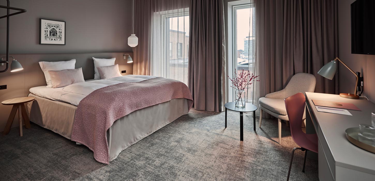 I venstre side står en dobbelt seng med lyserødt sengetæppe og pyntepuder. Til højre i rummet står et skrivebord og en lyserød skrivebordsstol. Foran et vindue står en lænestol og et sofabord med vase og blomst.