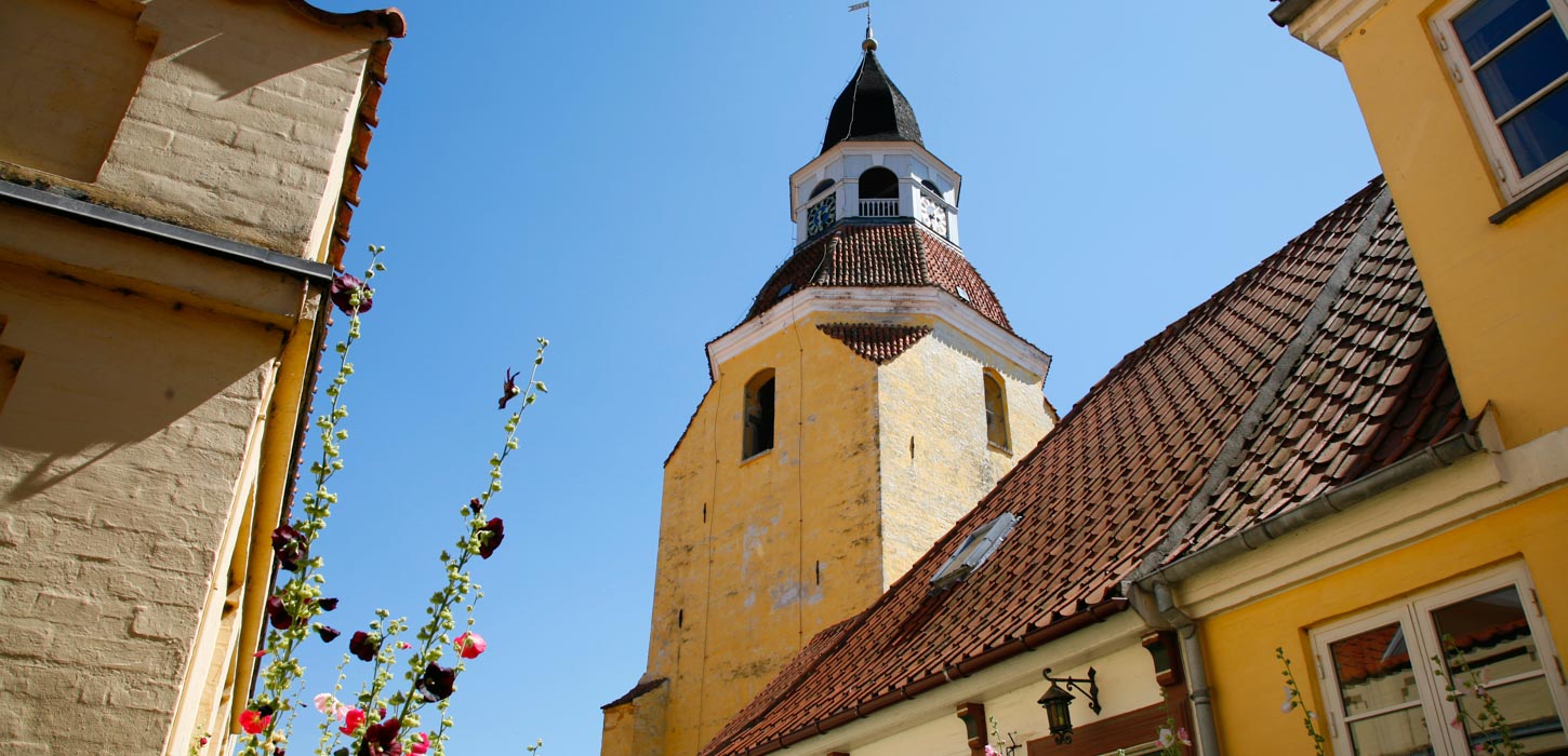 Det gule klokketårn med de røde tage i Faaborg set fra en skæv vinkel nedefra. Omkranset af blå himmel og stokroser.