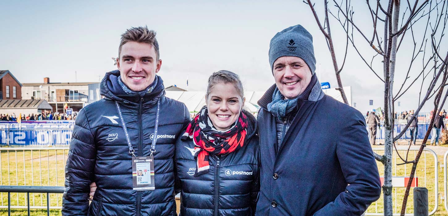 Kronprins Frederik står og smiller til kameraet sammen med en mand og en kvinde. I baggrunden er en grøn græsplæne, og bannere. I baggrunden ses opsætningen til løbet "Royal Run". 