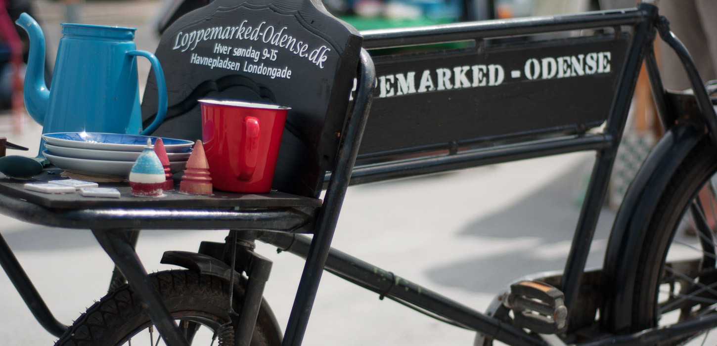 En nært billede af en sort gammel cykel. Cyklen har et lille lad foran med teksten "Loppemarked-Odense.dk" skrevet på. På ladet står en blå kande, par blå tallerkner og en rød kop.